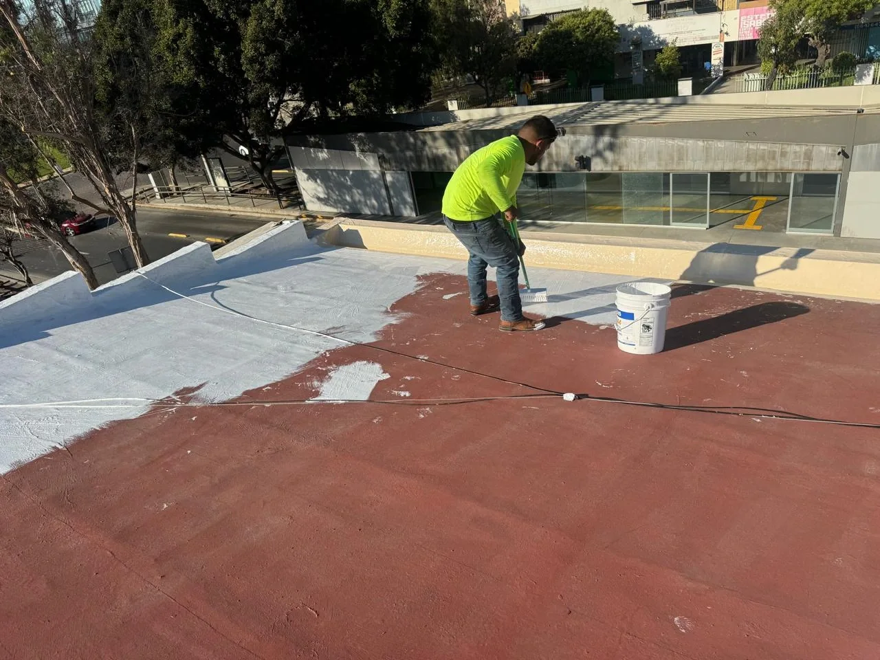 Trabajador extendiendo recubrimiento blanco sobre una azotea roja con una herramienta de mango largo.