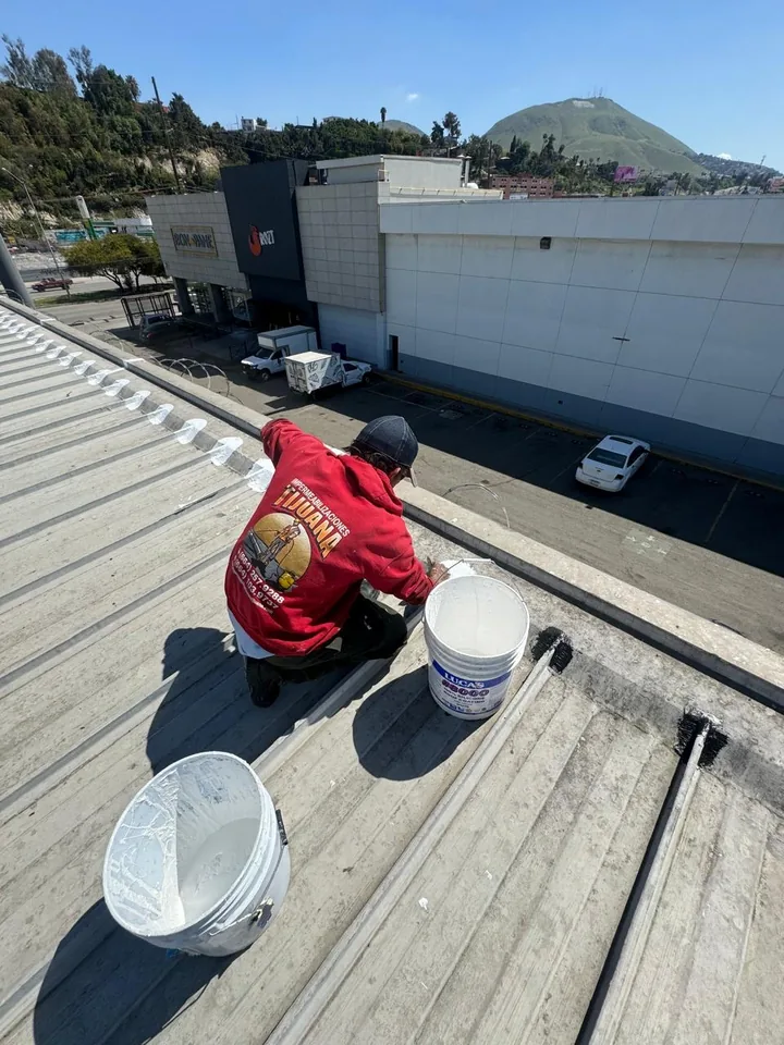 Trabajador aplicando recubrimiento blanco en el borde de una cubierta de lámina junto a una cubeta.