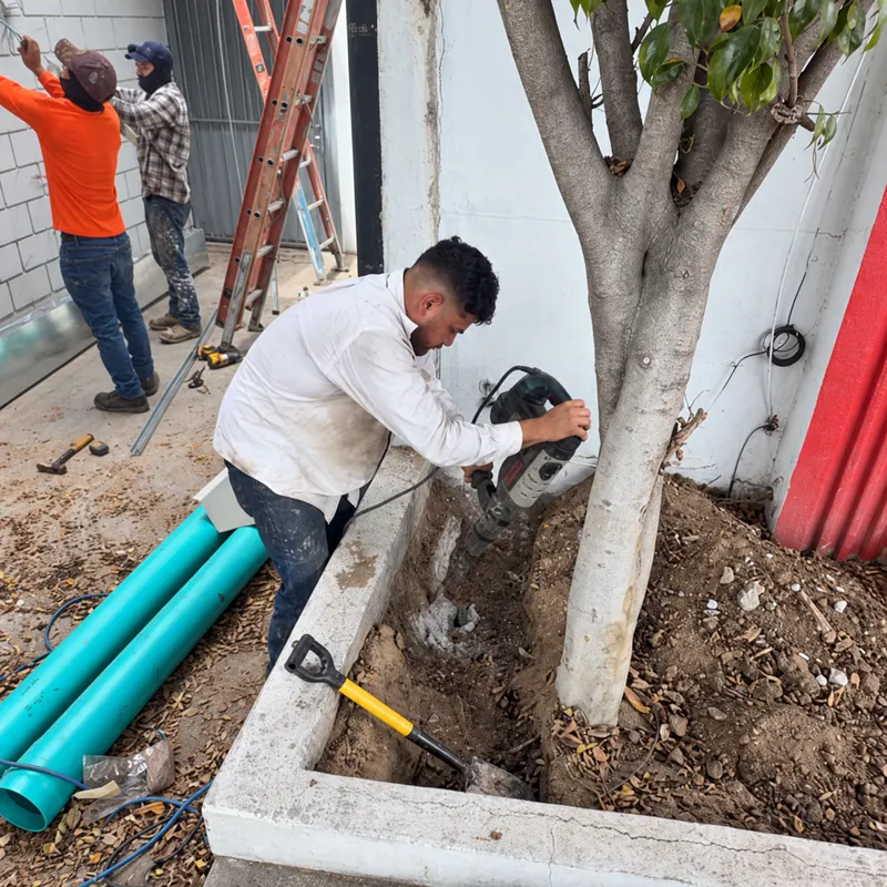 Trabajador rompe el concreto de una jardinera con rotomartillo para instalar tubería de drenaje, con dos compañeros al fondo