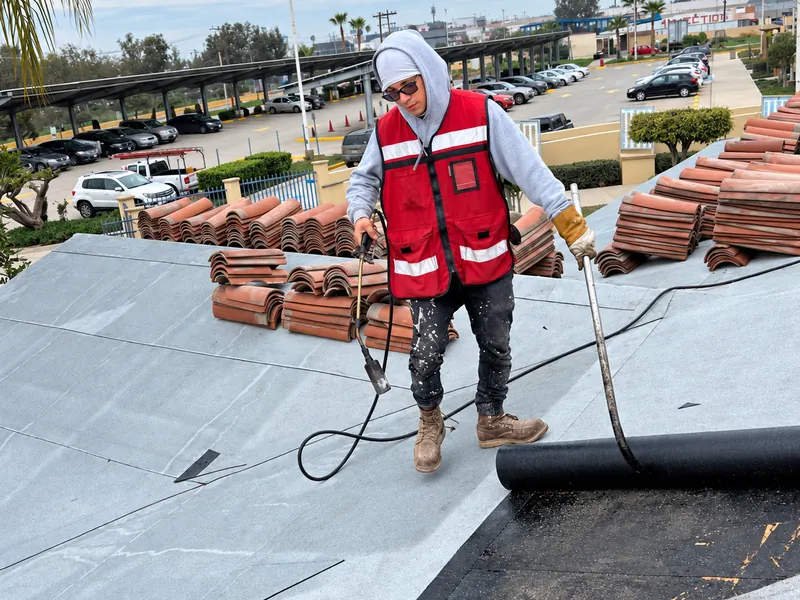 Trabajador con chaleco rojo manipula un rollo de membrana asfáltica junto a tejas apiladas