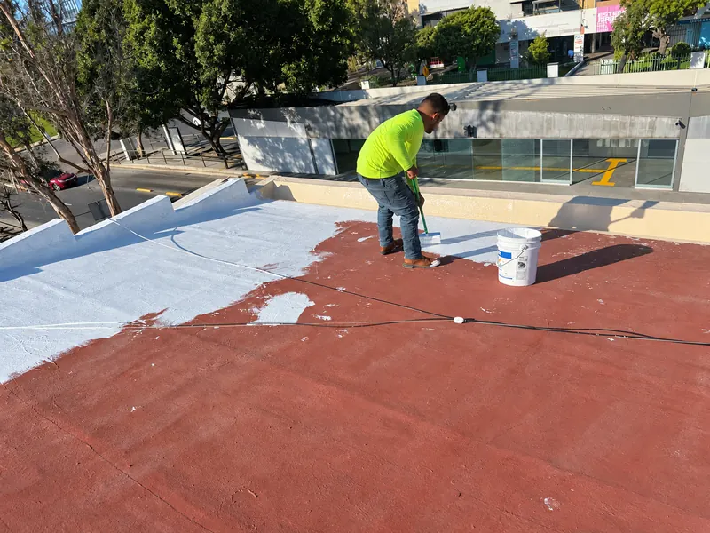 Trabajador extiende recubrimiento blanco con cepillo de mango largo sobre una azotea roja